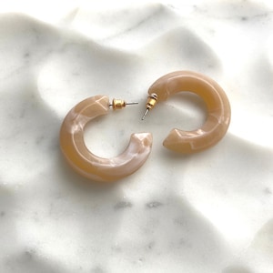May include: A pair of chunky, light brown hoop earrings with a marbled effect. The earrings have gold-colored posts and are set against a white, textured surface. These earrings are a stylish accessory.