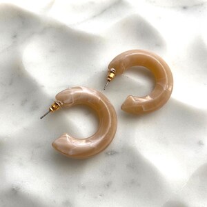 May include: A pair of light brown hoop earrings with gold-colored posts. The earrings are a marbled, translucent material, and are resting on a white, textured surface. The earrings are a simple, yet elegant accessory.
