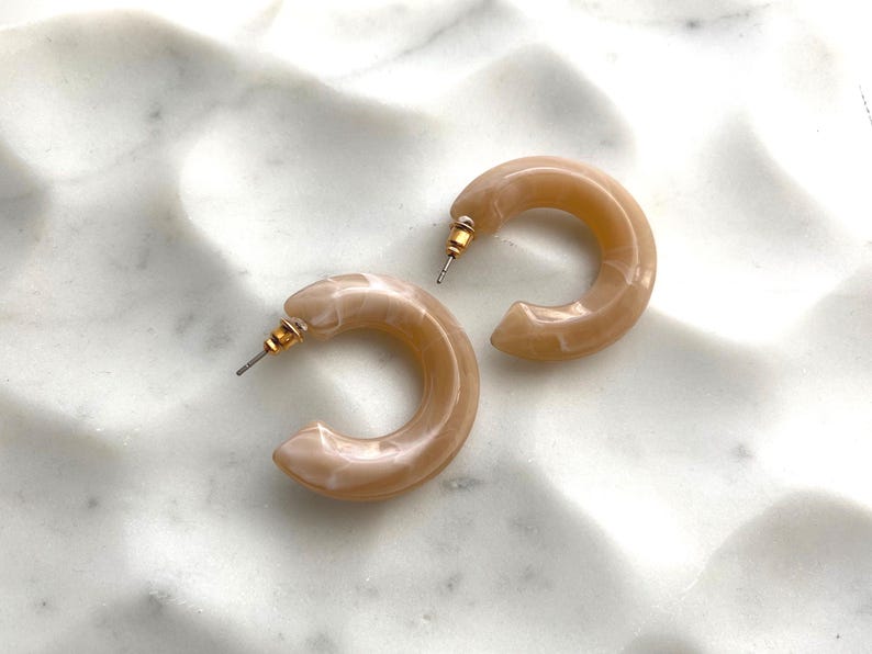May include: A pair of light brown hoop earrings with gold-colored posts. The earrings are a marbled, translucent material, and are resting on a white, textured surface. The earrings are a simple, yet elegant accessory.