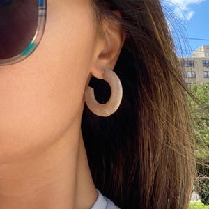 May include: A close-up of a person wearing light brown hoop earrings. The earrings are a thick, rounded shape. The person is also wearing sunglasses with a black frame and a blue shirt. The background includes a blue sky and a building.