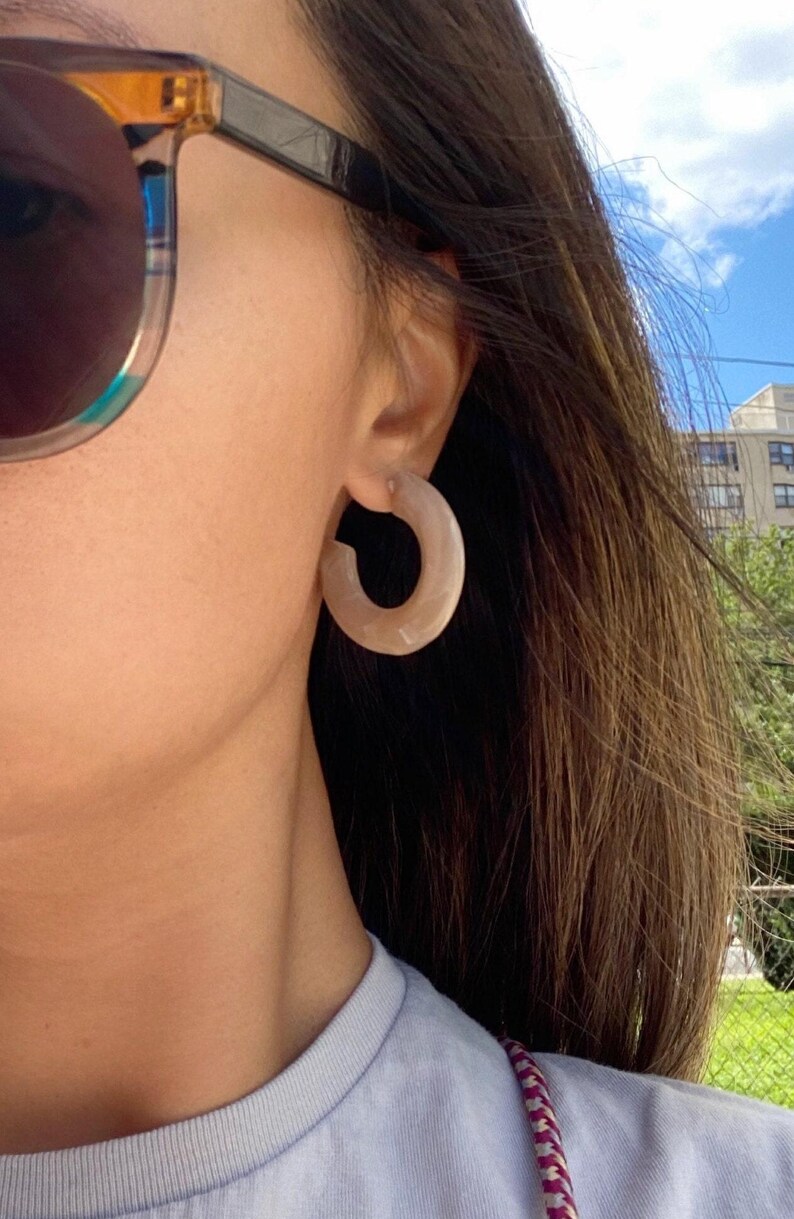 May include: A close-up of a person wearing light brown hoop earrings. The earrings are a thick, rounded shape. The person is also wearing sunglasses with a black frame and a blue shirt. The background includes a blue sky and a building.