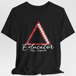 May include: Black t-shirt with a red triangle design featuring white pearls around the perimeter. The text "Educator" is written in cursive below the triangle, followed by "Mrs. Carpenter" in a smaller font.