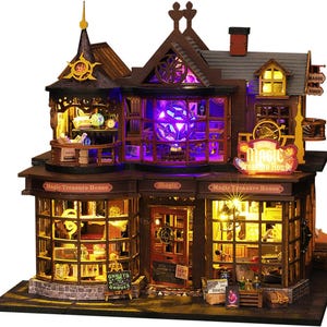 Op de afbeelding: Een houten modelbouwpakket van een magische winkel met een donkerbruine buitenkant en een bord met de tekst "Magic Treasure House". De winkel heeft drie ramen met verschillende items erin, waaronder een ketel, een kristallen bol en een spreukenboek. De winkel is verlicht met paarse en gele lampen.