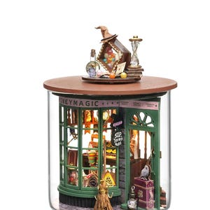 May include: A miniature glass jar diorama of a shop called "Honeydukes", a fictional candy shop from the Harry Potter series. The shop has a green exterior with a sign that reads "Shop That's Filled With Magic". The diorama includes a variety of details, such as a broom, a cauldron, and a stack of books.