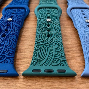 May include: A collection of silicone watch bands in various colors including black, dark blue, teal, and light blue. Each band features an intricate, embossed paisley-style pattern across the surface, a metal pin closure, and black connectors.
