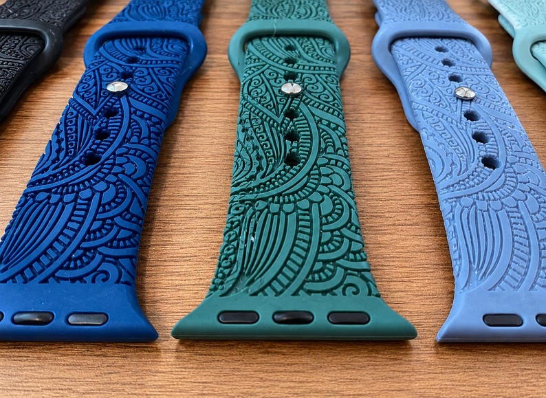May include: A collection of silicone watch bands in various colors including black, dark blue, teal, and light blue. Each band features an intricate, embossed paisley-style pattern across the surface, a metal pin closure, and black connectors.