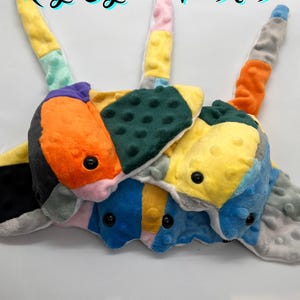 May include: A collection of colorful, patchwork plush toys. Each plush features a unique combination of vibrant colors like orange, yellow, blue, and green, with black button eyes. The text "Ray-Cycled Plushies" is displayed at the top.