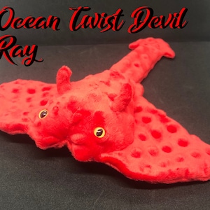 May include: A red plush devil ray toy with yellow eyes and a textured surface. The toy has the words "Ocean Twist Devil Ray" in black and red script across the top.