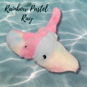 Peut inclure: Deux raies manta en peluche aux couleurs pastel, une rose et une blanche, avec des yeux noirs. Le texte "Rainbow Pastel Ray" est écrit en cursive. Le fond est une texture bleu clair, semblable à de l'eau.