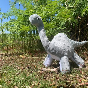 May include: A grey plush dinosaur toy with a long neck and a spotted back, standing in a grassy area with ferns in the background.