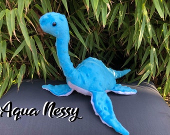 May include: A plush, turquoise Nessie toy with a long neck and small flippers. The toy has a textured body and white accents. The text "Aqua Nessy" is written in a cursive font at the bottom of the image.