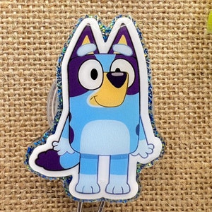 May include: A blue and purple cartoon dog with white eyes and a yellow nose. The dog is standing on a clear background with glitter around the edges. The dog is attached to a silver badge holder.