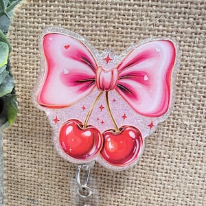 May include: A pink bow-shaped badge reel with a pair of red cherries. The bow has a glittery finish and small heart details. The cherries are glossy and attached by gold stems. A clear plastic retractable reel is attached.