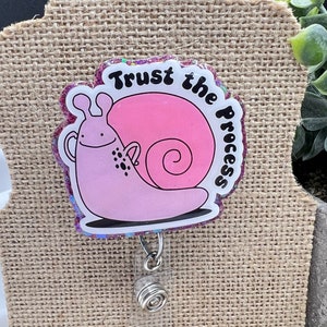May include: Pink glitter snail with black eyes and spots, with the text "Trust the Process" in black lettering. The snail is attached to a clear plastic retractable badge holder with a silver metal spring.