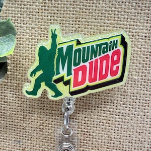 May include: A yellow and green badge reel with the words "Mountain Dude" in green and red. A green silhouette of a bigfoot figure is on the left. The badge reel has a clear plastic strap and a silver metal clip.
