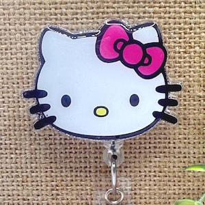 May include: A white and pink Hello Kitty retractable badge reel. The badge reel features a clear plastic base with a silver metal clip and a retractable cord. The Hello Kitty design has a pink bow and black whiskers.