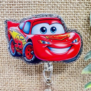 May include: A red Lightning McQueen badge reel with a glittery silver border. The cartoon car has blue eyes, a smiling mouth, and the number 95 on the side. The badge reel has a retractable cord and a silver clip.