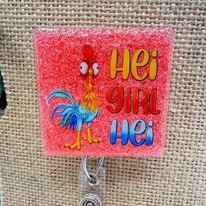 May include: A square, glittery red badge reel featuring a cartoon rooster with the words "Hei Girl Hei" in yellow, red, and blue. The rooster has orange, blue, and yellow feathers. The reel has a clear plastic strap and a silver metal clip.