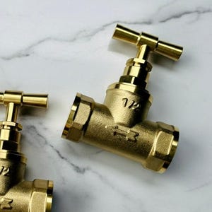 Pode incluir: Duas conexões de tubulação em forma de T de latão com acabamento polido. Cada conexão tem uma válvula com alça em T e é marcada com "1/2" e um símbolo de seta. As conexões estão em um fundo de mármore branco.