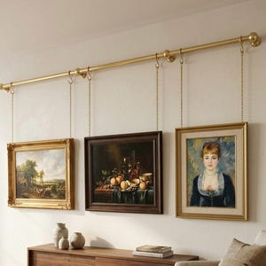 May include: Three framed paintings hanging from a gold-colored rod with chains. The paintings feature landscapes, still life, and a portrait. The frames are gold and dark brown. The paintings are displayed above a wooden cabinet.