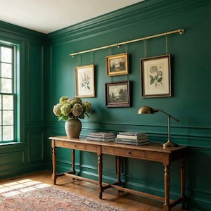 May include: A room with dark green walls, a wooden console table, and framed botanical prints. A brass rod holds the artwork. A vase of flowers and a brass desk lamp sit on the table. The floor is hardwood with a patterned rug.