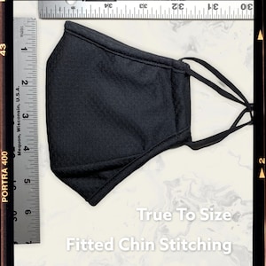 Peut inclure: Un masque facial noir avec des boucles d'oreille r&eacute;glables et une couture de menton ajust&eacute;e. Le masque est pos&eacute; &agrave; plat sur une surface blanche avec une r&egrave;gle &agrave; c&ocirc;t&eacute;. Le texte "True To Size" et "Fitted Chin Stitching" est imprim&eacute; sous le masque.