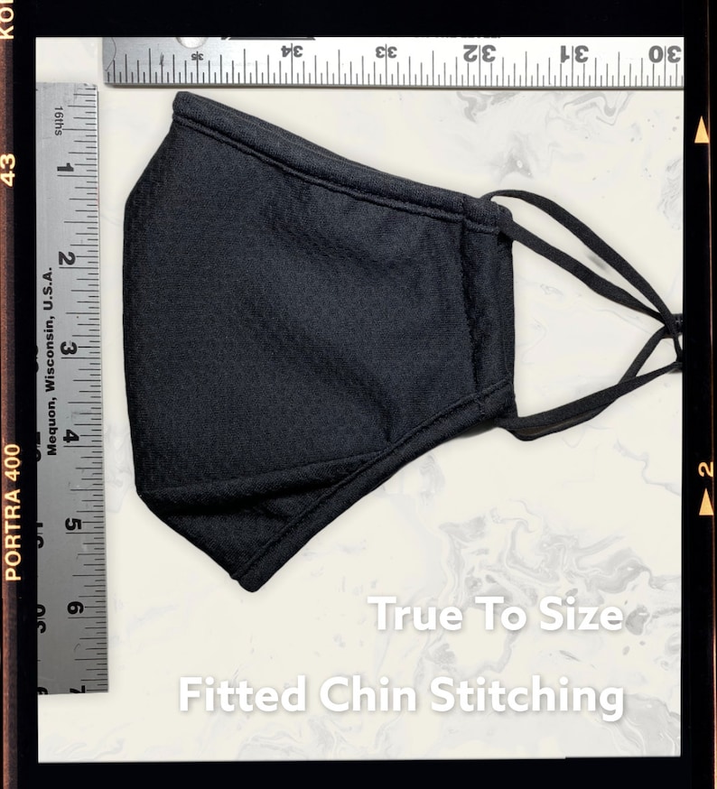 Peut inclure: Un masque facial noir avec des boucles d'oreille r&eacute;glables et une couture de menton ajust&eacute;e. Le masque est pos&eacute; &agrave; plat sur une surface blanche avec une r&egrave;gle &agrave; c&ocirc;t&eacute;. Le texte "True To Size" et "Fitted Chin Stitching" est imprim&eacute; sous le masque.
