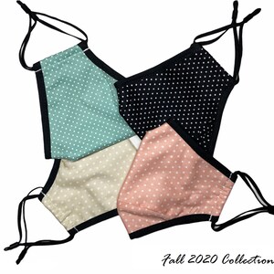 May include: Four fabric face masks with polka dot patterns in black, pink, beige, and green. The masks have black elastic straps and a black trim around the edges. The text "Fall 2020 Collection" is written below the masks.