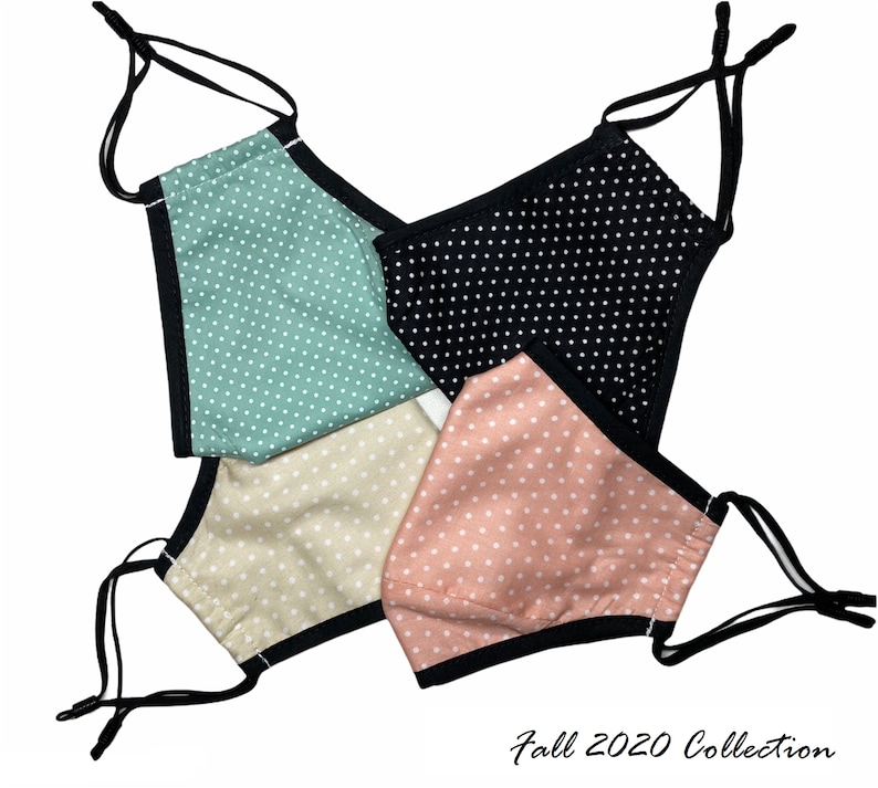 May include: Four fabric face masks with polka dot patterns in black, pink, beige, and green. The masks have black elastic straps and a black trim around the edges. The text "Fall 2020 Collection" is written below the masks.