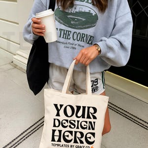 May include: A light gray sweatshirt with a green graphic and the text "THE CORNER". A cream-colored tote bag with the text "YOUR DESIGN HERE" is held in front. The bag is made of canvas. The person is wearing white shorts and sneakers.