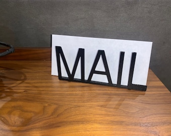 May include: Black metal mail holder with the word "MAIL" in large letters. The holder is sitting on a wooden surface.