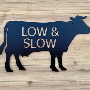 Pode incluir: Silhueta de vaca de metal preto com as palavras "LOW & SLOW" cortadas da silhueta.