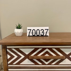 May include: A white wooden sign with the number 70037 in black lettering, sitting on a wooden dresser with a geometric pattern.
