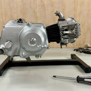 May include: A silver motorcycle engine with black accents is mounted on a black metal stand. The engine has a silver casing with a black cylinder head and a silver spark plug. The engine is on a white surface with a screwdriver lying next to it.
