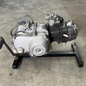 May include: A silver and black motorcycle engine mounted on a black metal stand. The engine features a prominent kick-start lever on the left side, a carburetor attached to the top, and a spark plug visible on the side of the cylinder head.