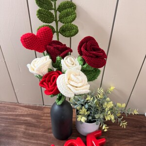 Puede incluir: Un ramo de crochet hecho a mano con rosas rojas y crema, un corazón rojo y hojas verdes. El ramo está en un jarrón negro, con un letrero de cerámica roja "LOVE" delante.
