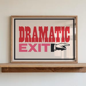May include: A framed rectangular wall art piece featuring the red text DRAMATIC and the pink text EXIT, accompanied by a black and white graphic of a hand pointing to the right. The print is enclosed in a light-coloured wooden frame.