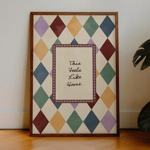 May include: A framed artwork with a diamond pattern in shades of purple, burgundy, yellow, green, and blue. The artwork features the text "This Feels Like Home" in a central rectangular frame with a decorative border. The frame is a dark wood.