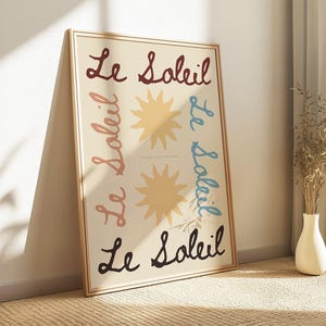 May include: A framed art print featuring the words "Le Soleil" in a cursive font, repeated in different colours. The print also includes two yellow sun illustrations. The artwork is leaning against a wall.