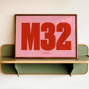 May include: A rectangular framed print featuring a light pink background with large, bold, red lettering that reads M32. Below the main text, a smaller, rounded rectangular outline contains the word STRETFORD in red capital letters.