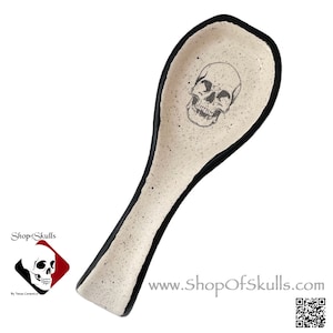 May include: A ceramic spoon rest with a skull design. The spoon rest has a speckled cream-colored surface with a black rim. The skull is centered on the spoon rest. The website www.ShopOfSkulls.com is visible.