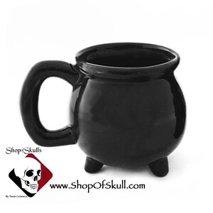 May include: A glossy black ceramic mug shaped like a cauldron, featuring a rounded body, a curved handle, and three small feet. The mug has the text "Shop of Skulls" and "www.shopofskull.com" printed on it.