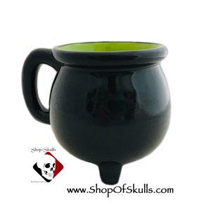 May include: Black ceramic cauldron mug with a green interior and a handle. The mug has three small feet and is shaped like a cauldron. The mug is a Halloween themed item.