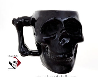 May include: A glossy black ceramic mug shaped like a human skull with a handle designed to resemble a human arm bone. The mug features the text ShopOfSkulls and the website address www.ShopOfSkulls.com printed on the lower portion of the image.