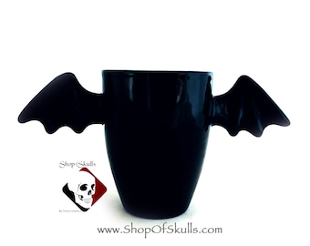 May include: Black ceramic mug with bat-shaped handles. The mug is decorated with a skull logo and the text "Shop of Skulls" and "www.shopofskulls.com".