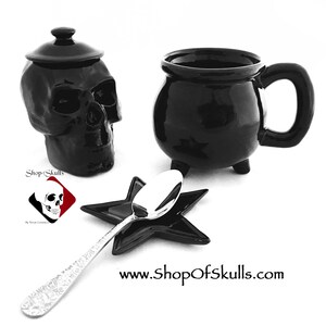 May include: A black skull-shaped container with a lid, a black cauldron-shaped mug, a star-shaped spoon rest, and a silver spoon. The items are arranged on a white surface. The website address www.shopofskulls.com is visible.