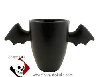 May include: A matte black ceramic mug featuring two sculpted bat wings extending from the sides. The mug has a smooth, solid black finish. The logo for Shop of Skulls by Texas Ceramics and the website www.ShopOfSkulls.com are printed below.