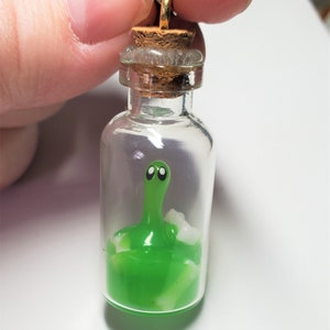 以下が含まれることがあります： 緑色の粘着性のある生き物が閉じ込められた小さなガラス瓶のチャーム。生き物は黒い目を2つと、粘着性の中に白い渦巻き模様があります。瓶にはコルク栓と金色のチェーンが付いています。