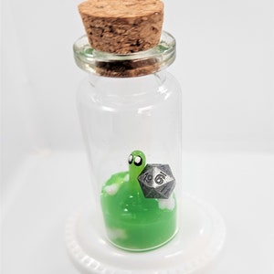 May include: A small glass bottle with a cork stopper contains a green slime with a white cloud and a silver metal dice with the number 16 on it. The bottle is sitting on a white pedestal.