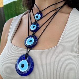 May include: Layered necklace with four blue evil eye pendants of different sizes. The pendants are round and have a white center with a black pupil. The necklace is made of black cord.